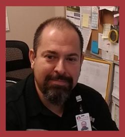This image is a headshot of the Director, Mr. Reyna. He is sitting at his desk. A large corkboard with pinned flyers and papers is behind him. He is wearing a black shirt, his badge, and he is smiling.