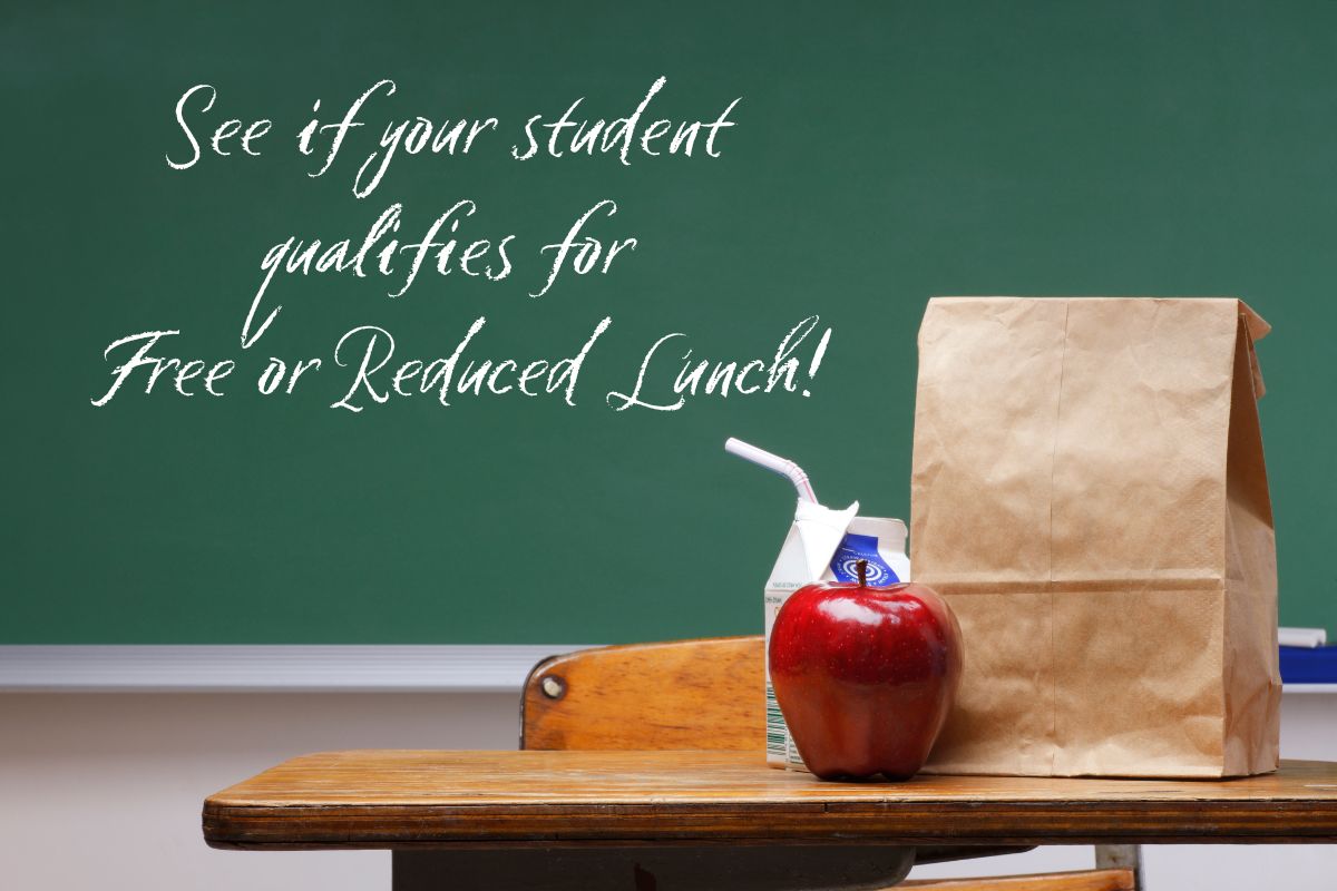 Brown paper lunch bag, milk carton, and apple on a wooden desk in the foreground. Chalkboard in the background with the text, "See if your student qualifies for Free or Reduced Lunch!"