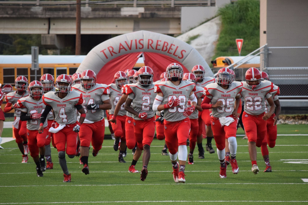 A group of 16+ student football players running onto the football field. They are wearing red and gray uniforms.